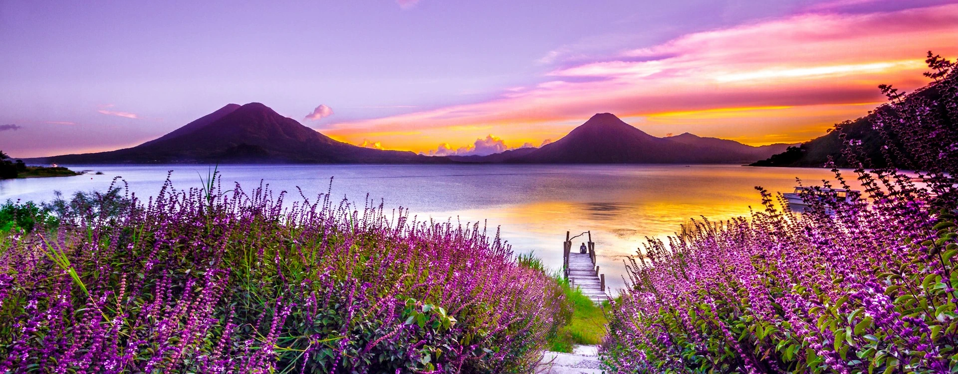 A beautiful sunset in Lake Atitlán in Guatemala, with a wooden dock surrounded by purple flowers, and the high peaks of the volcanoes seen in the distance