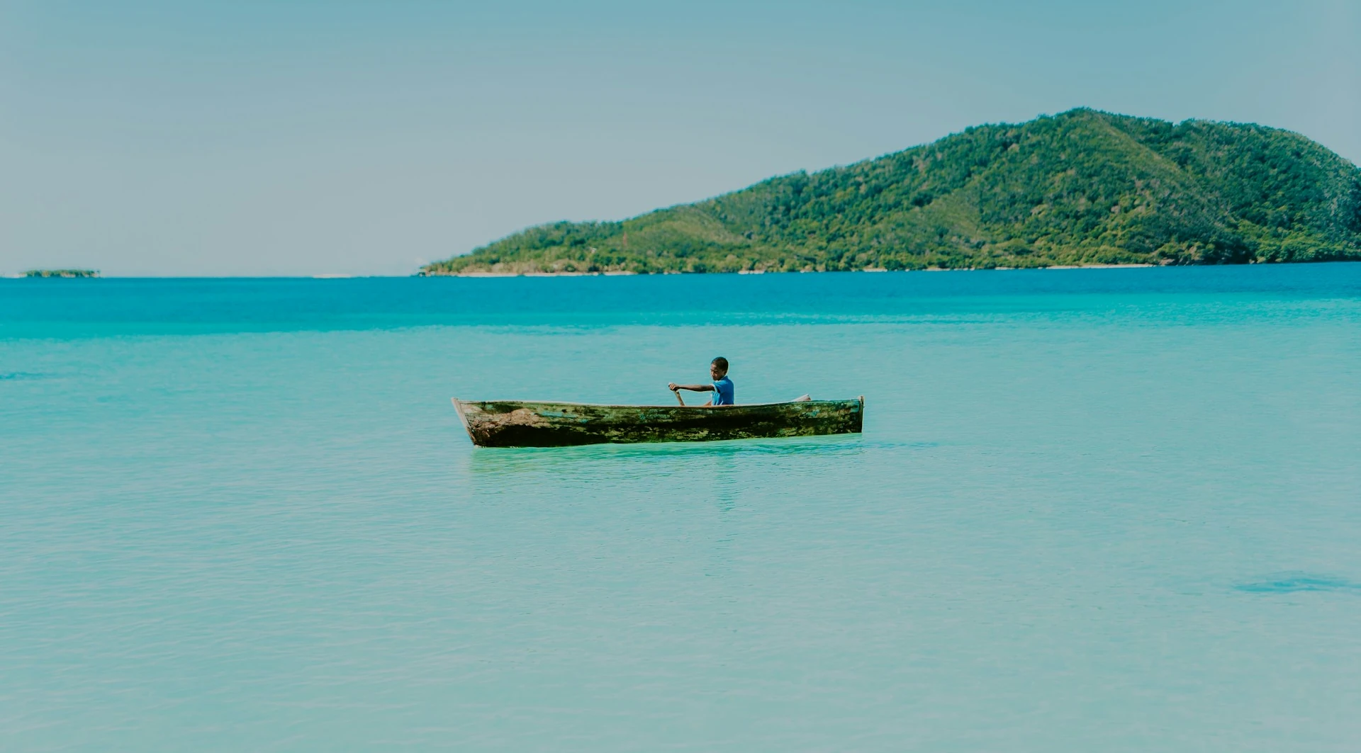 Person sailing a canoe on a calm sea with turquoise blue waters, with an island full of vegetation nearby
