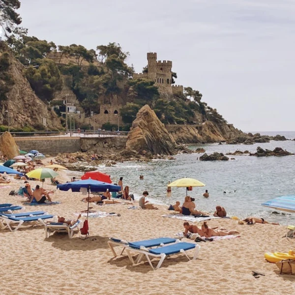 Golden sandy beach full of people sunbathing or swimming in its calm waters, with views of a castle on the cliffs
