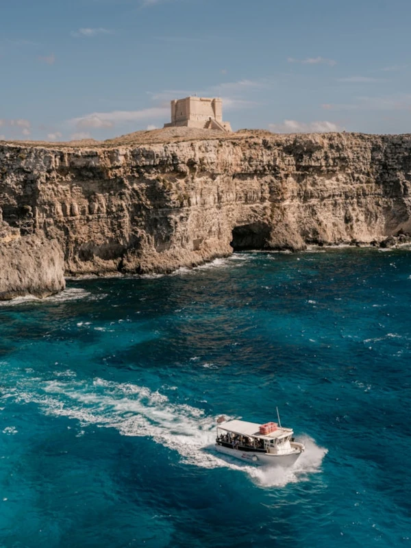 A boat navigating in the blue waters of Comino under the watch of Santa Marija Tower