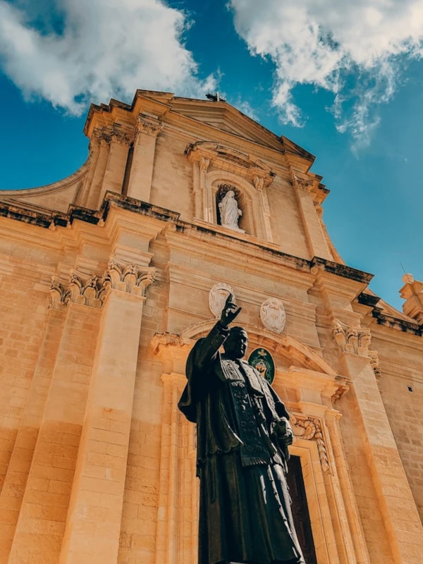 Facade of Victoria Cathedral in Gozo with an imposing statue in the foreground on a sunny day.