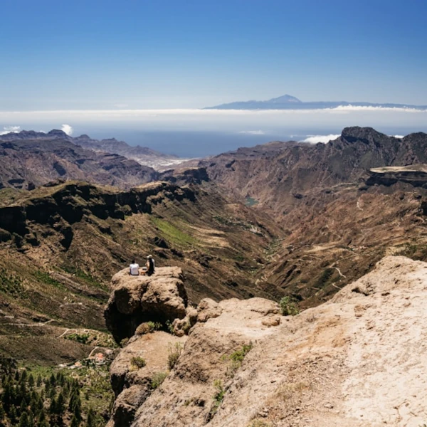 A couple sitting on a rock, looking down at the panoramic views of a deep valley of Gran Ganaria, with the Atlantic Ocean and Tenerife on the distance