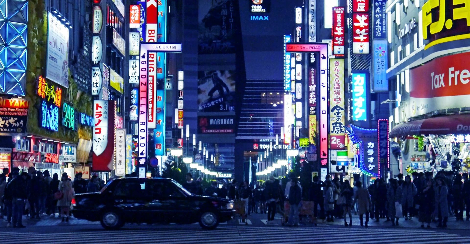 Una calle de Shinjuku iluminada con luces de neón, llena de luces y colores. La calle está llena de gente caminando y se ve un coche negro circulando.