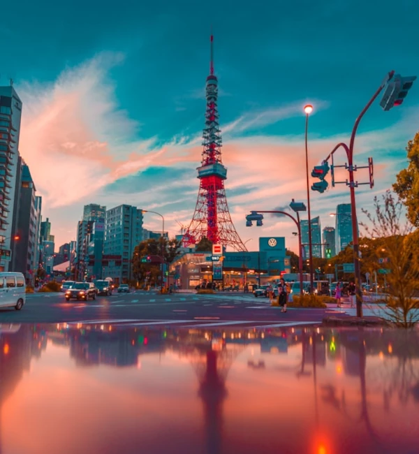 La Torre de Tokio entre edificios y semáforos durante el atardecer, con un charco en la calle que refleja las luces y la torre.