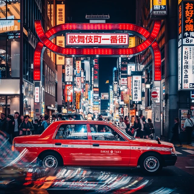 Un coche rojo circulando por una calle de Tokio. La calle está llena de luces de neón brillantes, carteles y gente caminando.