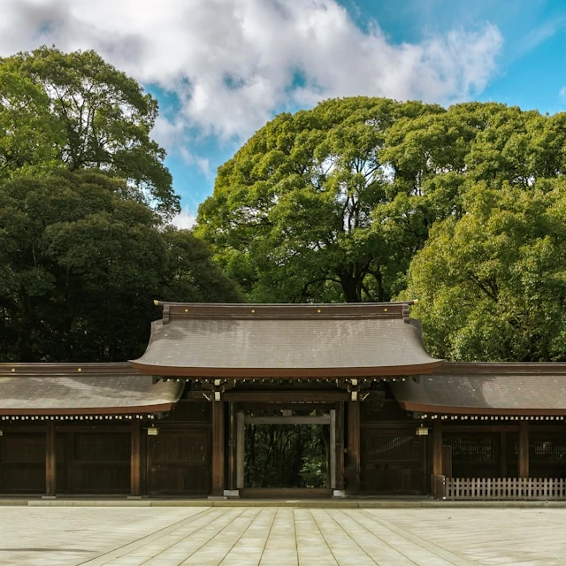 El patio vacío de un templo en Tokio, con árboles muy altos y frondosos al fondo, en un día soleado con algunas nubes en el cielo.