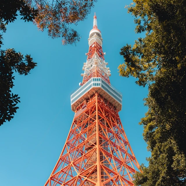 La parte superior de la Torre de Tokio, de color rojo y blanco, con árboles verdes que dan sombra a sus pies en un día claro y soleado.