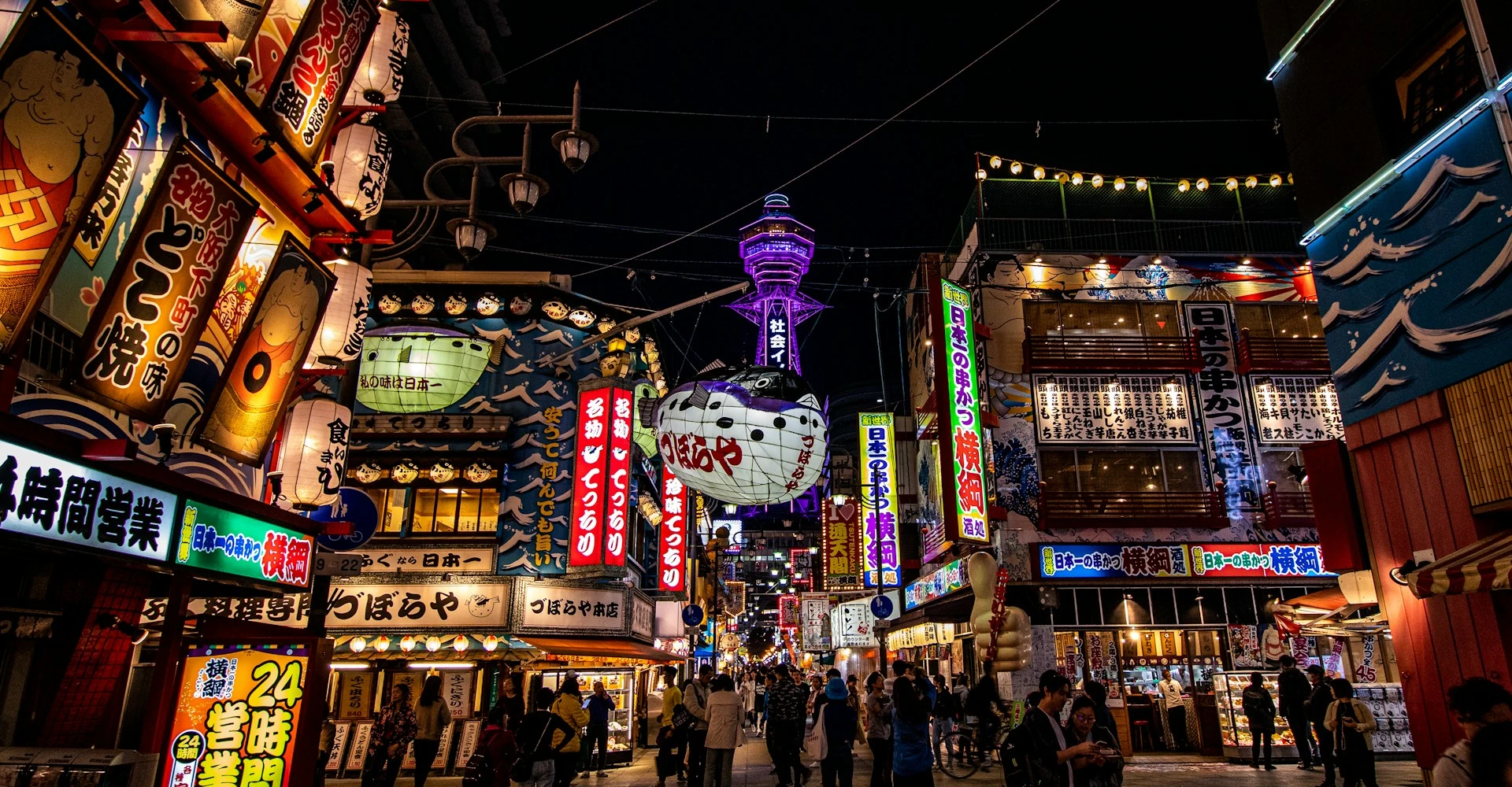 Vibrant urban market street with bustling activity and the iconic Tsutenkaku Tower in Osaka, Japan.