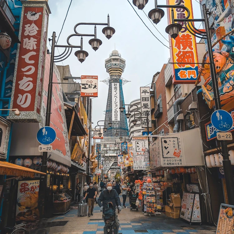 A market street with bustling activity and the iconic Tsutenkaku Tower in Osaka, Japan.