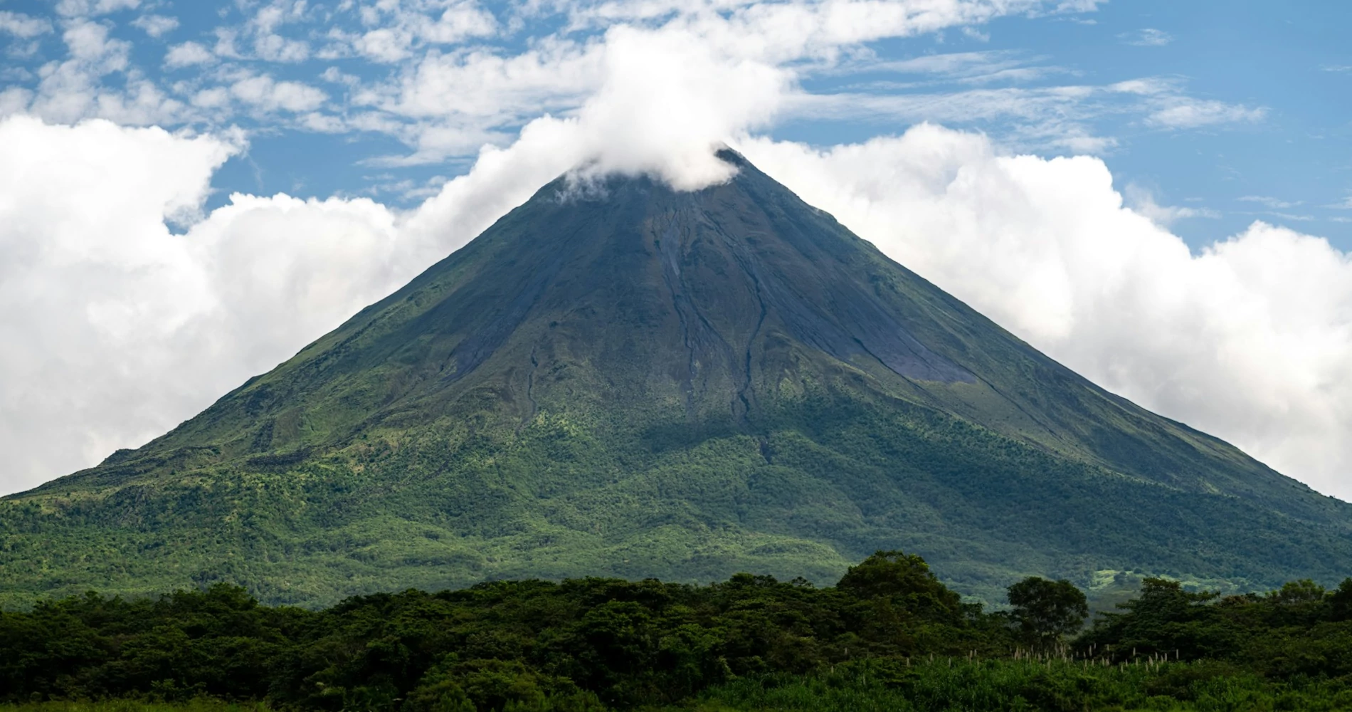 A very tall volcano towering over lush green vegetation beneath the blue sky and white clouds