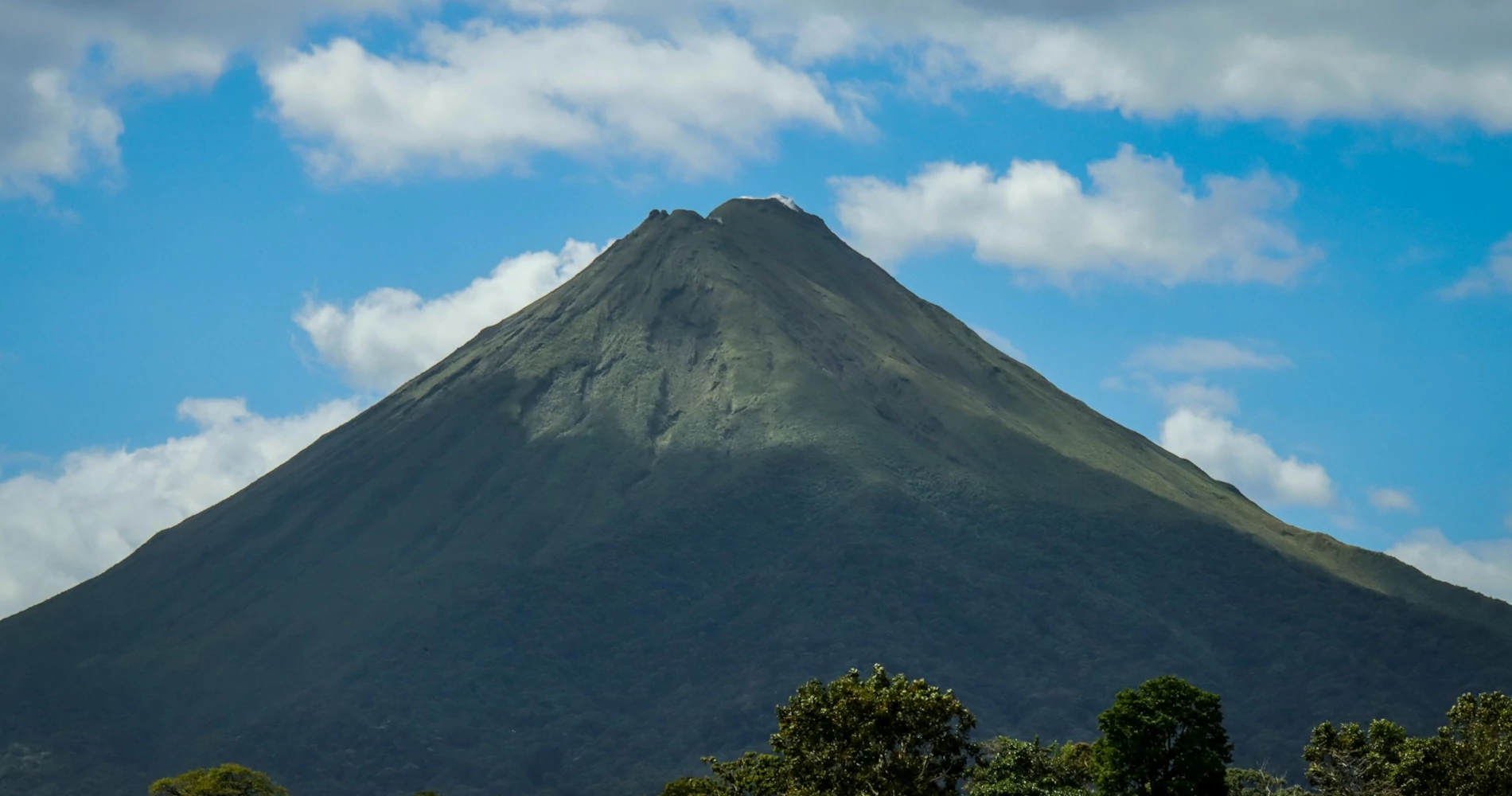 A very tall volcano towering over lush green vegetation beneath the blue sky and white clouds