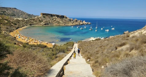View of Ghajn Tuffieha Beach in Malta from the stairs, with golden sand, turquoise waters, surrounding green cliffs, and people relaxing under umbrellas
